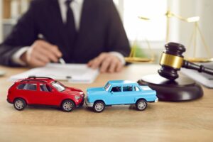 An image of two toy cars, one red and one blue, in a head-on collision. In the background, a person in a suit is writing, with a gavel and scales of justice on the desk, indicating a legal setting, likely related to a car accident case.